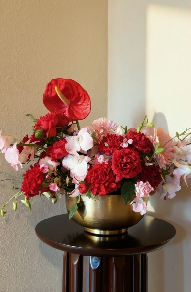 Red anthurium, carnations, and pink orchids arranged in a brass vase on a wooden pedestal
